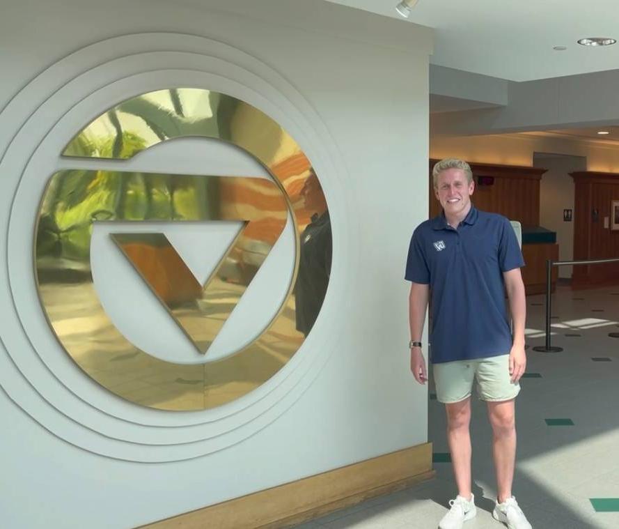 Male with blond hard, blue shirt and khaki shorts standing next to brass GVSU logo